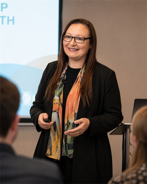 Jennifer Natale presenting to an audience, smiling mid-sentence with open hand gestures, holding a clicker, wearing a black blazer and colourful scarf.